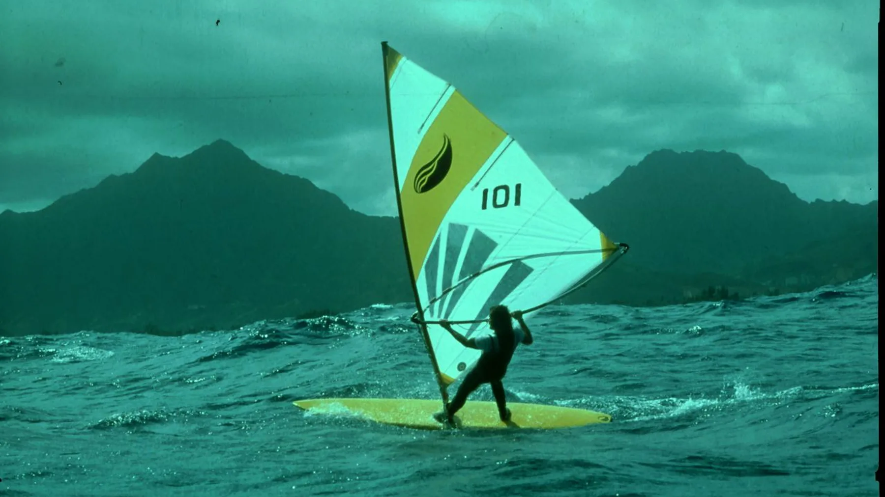 Windsurfer op een geel board met bergen op de achtergrond